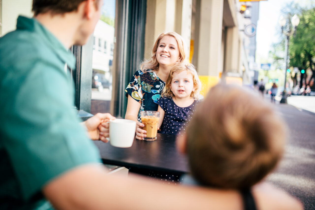 A happy young Caucasian family sits outside of a cafe / coffee shop enjoying hot drinks on a clear day. They have relaxed conversation, enjoying the time together.  The father has his arm around his son in the foreground, with the focus on the mother and daughter visible over their shoulders in the background.