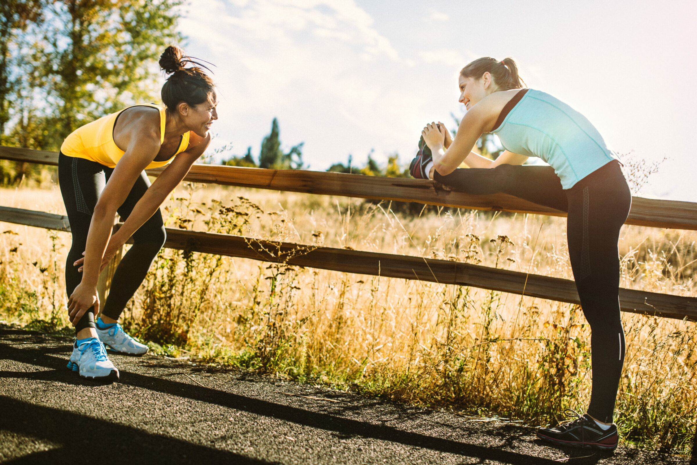 Two young adult women stretch out before running in a pristine and vast outdoor  area, the sun shining warm golden light on the scene.  Depicting trail running, healthy lifestyle and outdoor exercise in nature.  Horizontal image.