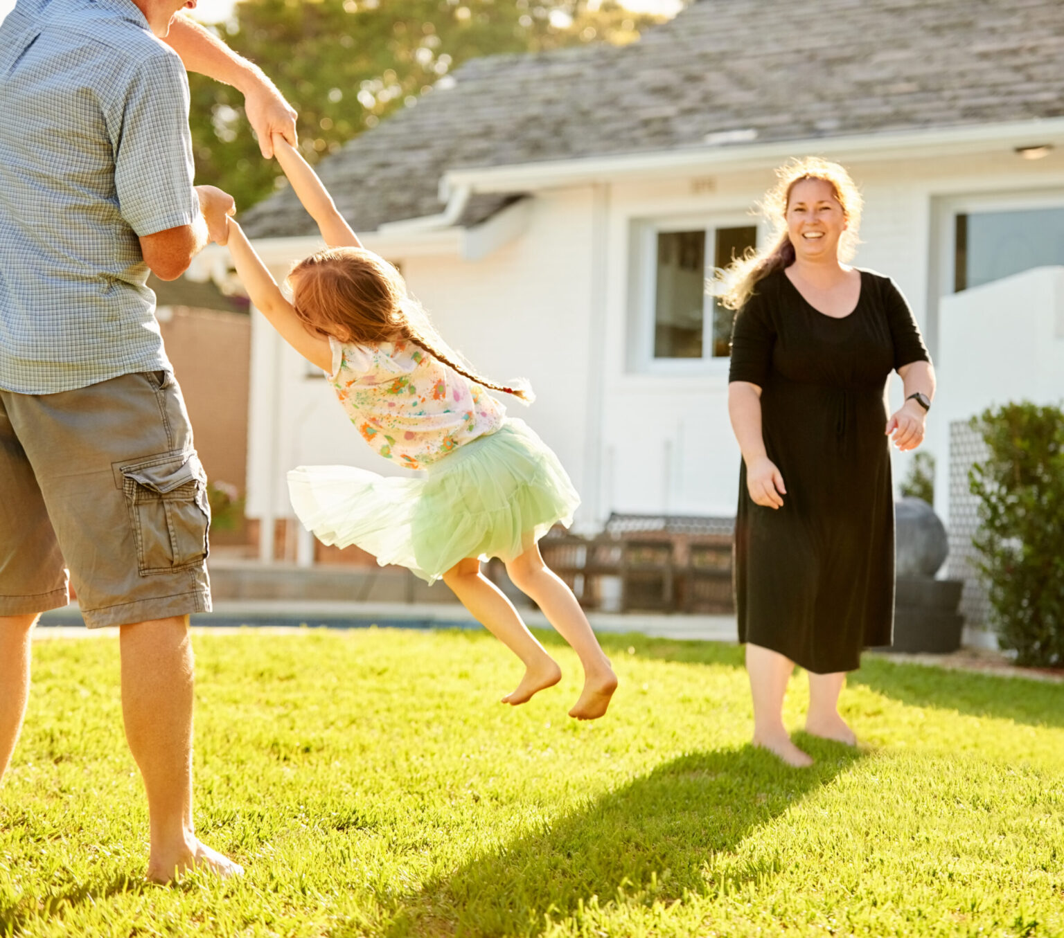 Father holding hands of her daughter and spinning her on backyard lawn on a summer day with mother standing in background